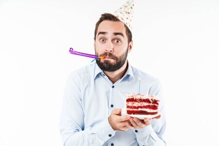 Image of positive man blowing party horn and holding birthday cake with candle isolated over white backgroundの写真素材