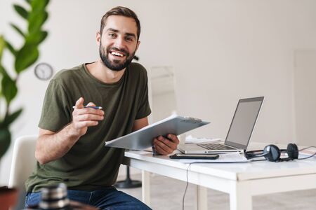 Image of smiling young man pointing finger at camera and holding clipboard while sitting at desk in bright officeの写真素材