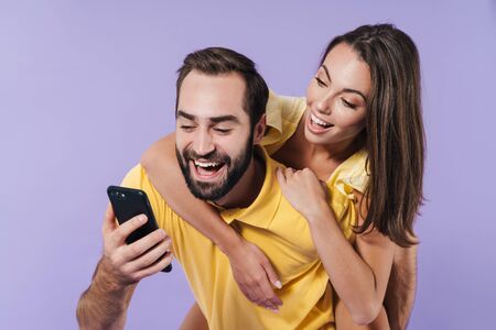Happy excited beautiful young couple wearing casual clothing standing isolated over violet background, piggyback ride, using mobile phoneの写真素材