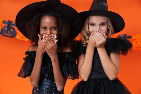 Image of cute multinational girls in black halloween costumes laughing and covering their mouths isolated over orange pumpkin wallの写真素材