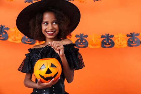 Image of joyful african american girl in black halloween costume smiling and holding toy pumpkin isolated over orange wallの写真素材