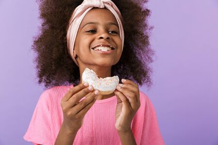 Image of a cute optimistic young african girl kid posing isolated over purple wall background eat donut.の写真素材