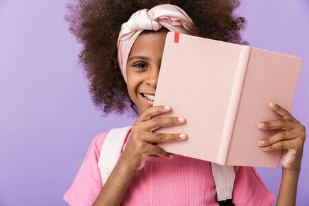 Image of a happy optimistic young african girl kid posing isolated over purple wall background covering face behind the bookの写真素材