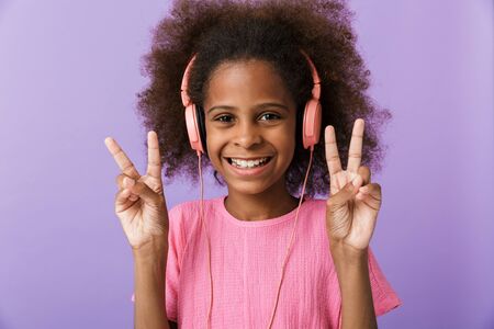 Image of a cheerful optimistic young african girl kid posing isolated over purple wall background listening music with headphones showing peace gesture.の写真素材
