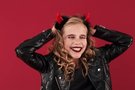 Picture of cheerful little girl devil in carnival halloween costume isolated over red wall...