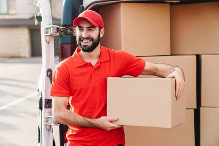Image of happy young delivery man in red uniform standing with parcel post box near car outdoorsの写真素材