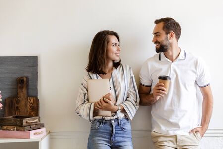 Smiling young couple standing at the wall in office, holding laptop computer and takeaway coffee cup