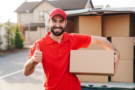 Image of smiling young delivery man in red uniform standing with parcel box and showing thumb up near car outdoorsの写真素材