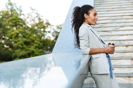Attractive smiling african business woman standing outdoors at the city, leaning on stairs, holding...