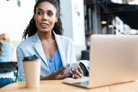 Attractive smiling young african businesswoman sitting at the cafe table outdoors, working on laptop computerの写真素材