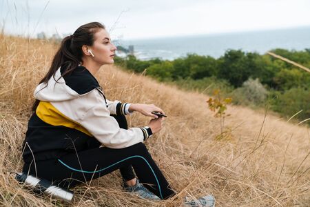 Photo of nice caucasian woman in sportswear using earpods and smartphone while sitting on dry grass at seashoreの写真素材