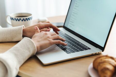 Cropped photo of young caucasian woman typing on laptop while having breakfast in cozy living room