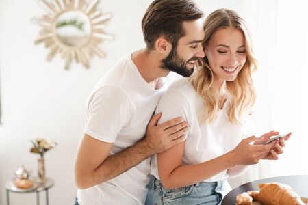 Happy beautiful young couple having breakfast at the kitchen table, using mobile phone, embracingの写真素材