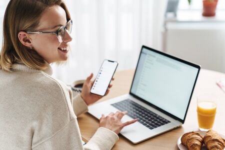Photo of happy caucasian woman typing on laptop and cellphone while having breakfast in cozy living...