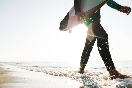 Cropped photo of caucasian young man in wetsuit holding surfboard while walking at sunny beachの写真素材