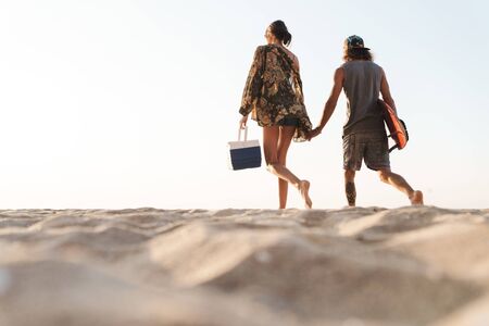 Photo from back of attractive couple holding surfboard and cooler bag while walking on sunny beach