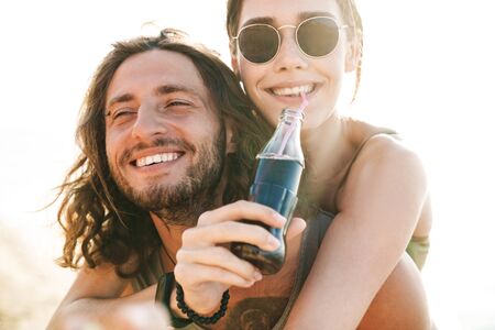 Photo closeup of happy caucasian man piggybacking nice woman who drinking soda while walking on sunnyの写真素材