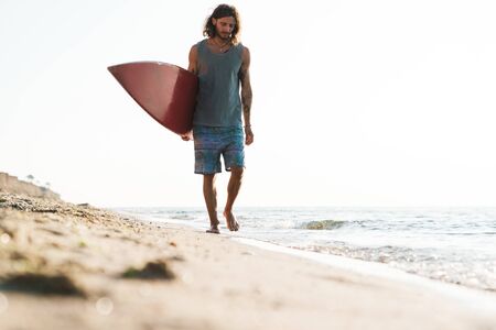 Photo of unshaven focused man in summer clothes holding surfboard while walking on sunny beach
