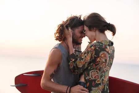 Happy young couple hugging while standing at the beach, holding surf boardの写真素材