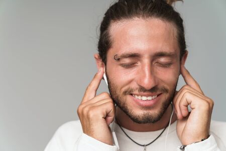 Close up of a smiling young man enjoys listening to music with earphones while standing isolated...