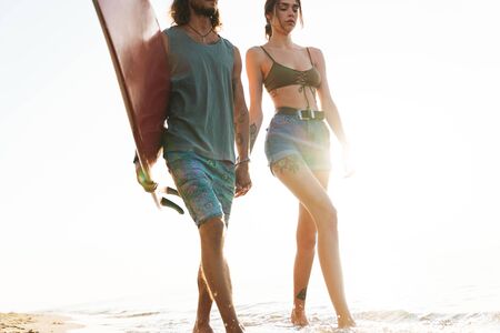 Photo of attractive serious couple with surfboard holding hands together while walking on sunny beachの写真素材