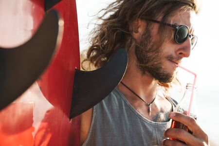 Photo of handsome young man in sunglasses drinking soda and holding surfboard on sunny beach