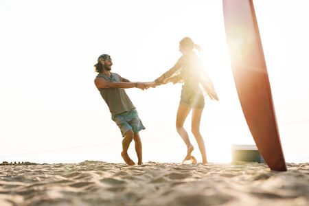 Photo of joyful caucasian couple with surfboard smiling and spinning while having picnic on sunny...