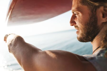 Photo of pleased confident man holding surfboard on his head and pointing finger at sea on sunny beachの写真素材