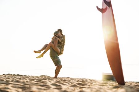 Photo of cheerful man with surfboard carrying slim woman in his arms while having picnic on sunny...