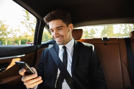 Image of young caucasian successful businesslike man in formal black suit using earbuds and...