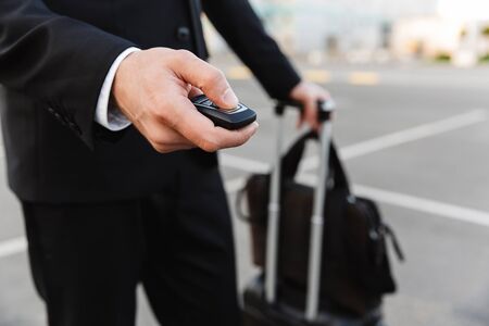 Image of young caucasian businesslike man in formal suit opening car with alarm remote key outdoors