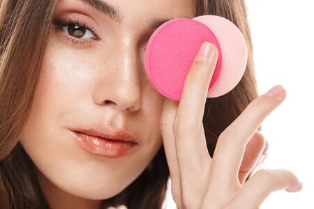 Portrait closeup of attractive young woman applying cosmetics with makeup sponges isolated over white backgroundの写真素材