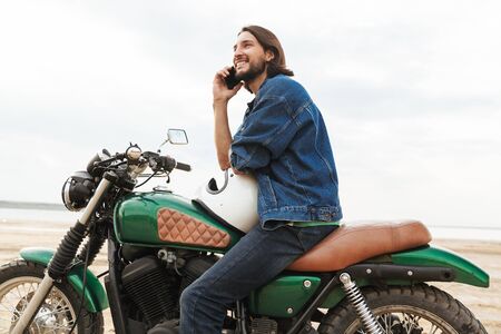 Picture of a smiling cheery man biker on his bike outdoors on a beach talking by mobile phone.