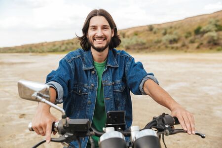Close up of a handsome happy young man wearing casual outfit sitting on a motocycle at the beachの写真素材