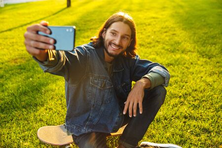 Handsome smiling young man spending time at the park, taking a selfie while sitting on grass with a skateboardの写真素材