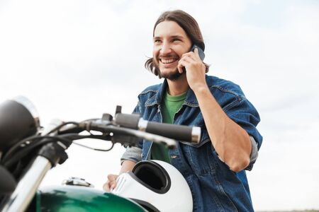 Close up of a handsome young man wearing casual outfit sitting on a motocycle at the beach, talking...