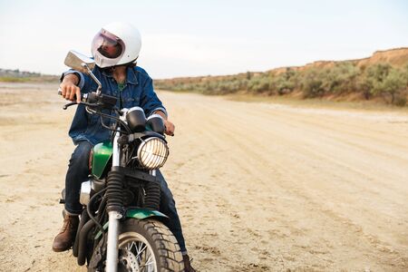 Handsome young man wearing casual outfit sitting on a motocycle at the beach, wearing a helmetの写真素材