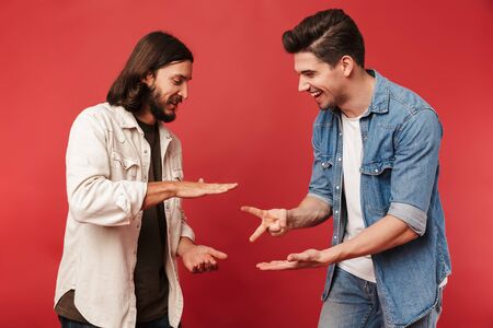 Photo of young smiling guys playing rock-paper-scissors game and laughing isolated over red...