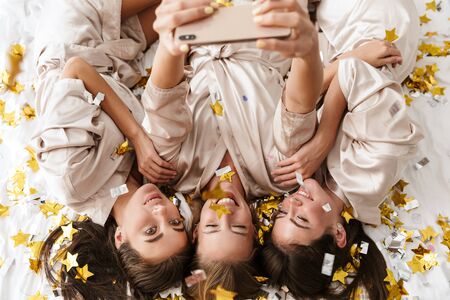 Image of a cheerful young girls women friends indoors on bed at the hen party at home lies over confetti take a selfie by mobile phone.の写真素材