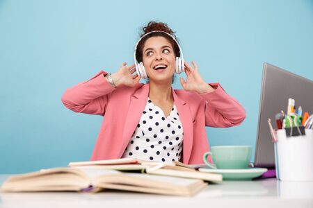 Image of smiling woman listening music with headphones while working at desk isolated over blue...