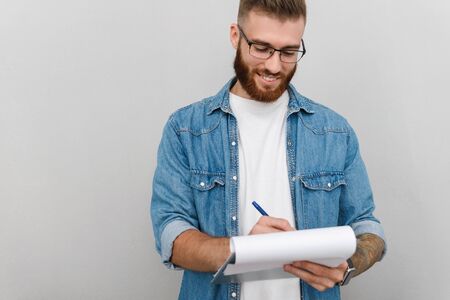 Image of cheerful handsome man in eyeglasses writing and holding clipboard isolated over gray backgroundの写真素材