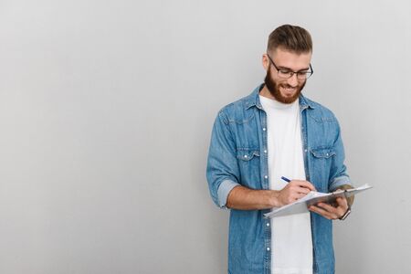 Image of cheerful handsome man in eyeglasses writing and holding clipboard isolated over gray backgroundの写真素材