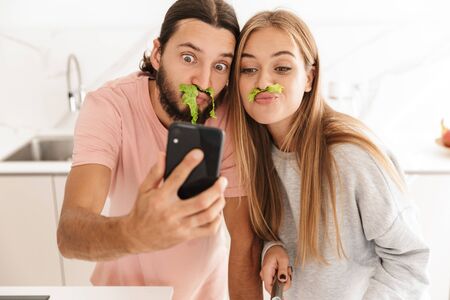 Photo of cute funny young loving couple at the kitchen indoors at home cooking together take a selfie by mobile phone having fun.の写真素材