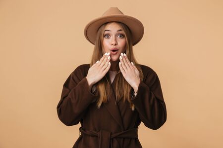 Image of young caucasian girl wearing hat and coat expressing wonder and raising hands at face...
