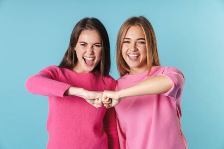 Photo of excited nice women in pink sweaters laughing and holding their fists together isolated over blue backgroundの写真素材