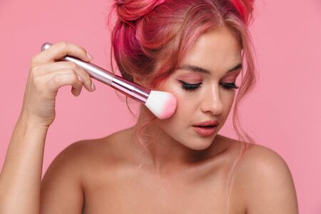 Portrait of caucasian shirtless woman with colorful hairstyle applying cosmetics with makeup brush isolated over pink backgroundの写真素材
