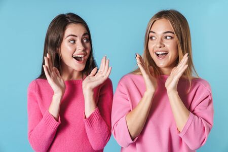 Two cheerful young girlfriends standing isolated over blue background, screaming, looking at each...