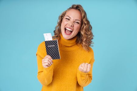 Cheerful lovely young girl wearing sweater standing isolated over blue background, showing passport...