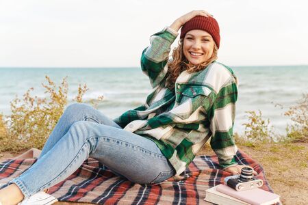 Image of joyful young caucasian woman wearing hat and plaid shirt sitting on blanket by seasideの写真素材