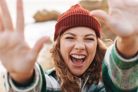 Image of joyful young caucasian woman wearing hat and plaid shirt reaching hands at camera while walking outdoorsの写真素材